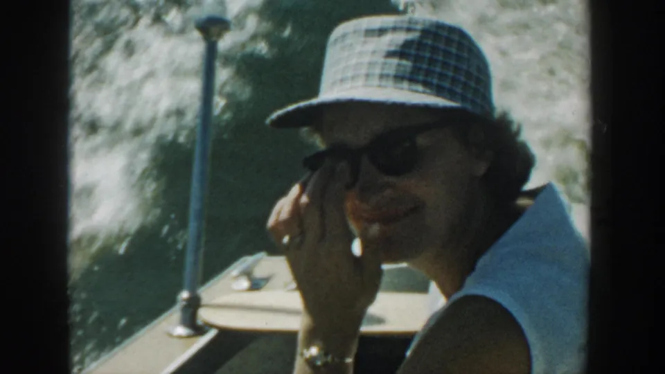 Adult woman on a Wisconsin lake boat in 1957, wearing dark sunglasses and a checkered sun hat while smiling toward the camera.