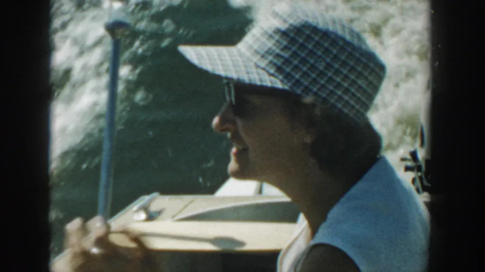 Profile of the woman in sunglasses and a checkered sun hat on a 1957 Wisconsin boating outing, with bright lake water behind her.