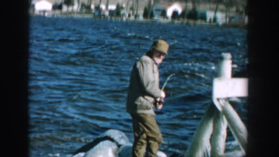 1957 Wisconsin 8mm frame showing a man in a light jacket and cap fishing near choppy lake water with houses across the shore.