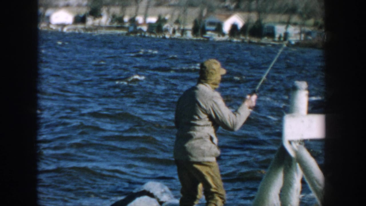1957 Wisconsin home-movie frame of a man fishing from a dock or boat edge, with rippled water and lakeside houses behind him.