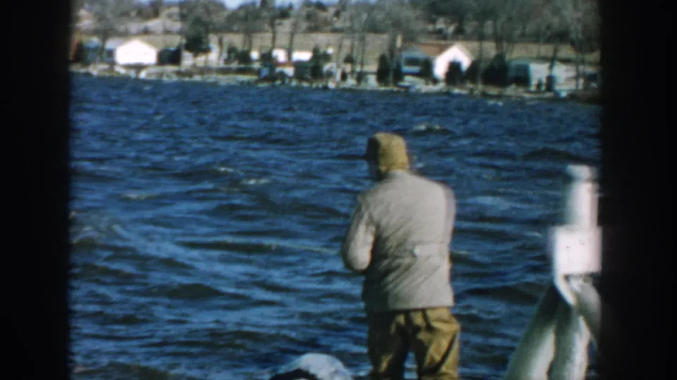 Rear view in 1957 Wisconsin 8mm footage of a man fishing over choppy lake water, framed by shoreline homes and dark film edges.