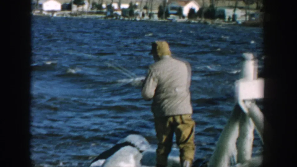 1957 Wisconsin frame showing a man fishing beside rocks and dock hardware, with wind-ruffled lake water and houses in the distance.