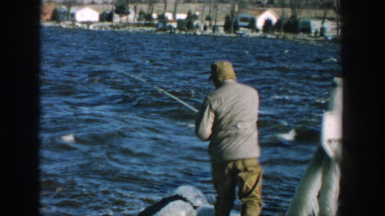 Adult in a light jacket fishes from a lakeside dock with houses across the water.