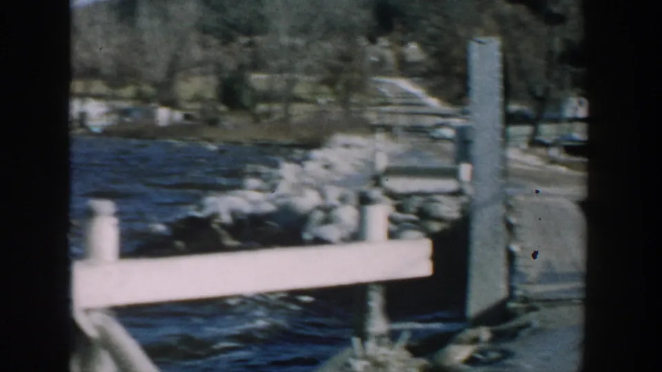 A 1957 Wisconsin home-movie frame shows the dock railing, rocks, lake water, and distant residential shoreline after the fisher leaves view.