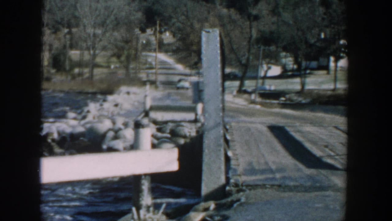 View from a wooden pier toward a road, shoreline rocks, water, bare trees, and distant cars.