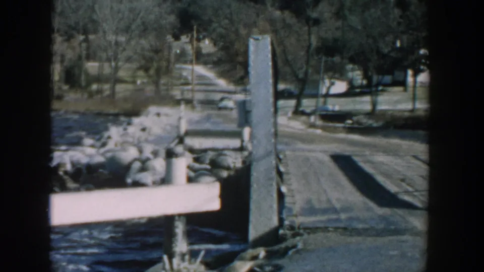 1957 Wisconsin home-movie frame showing a wooden pier beside blue water, shoreline rocks, a road, bare trees, and distant vehicles.