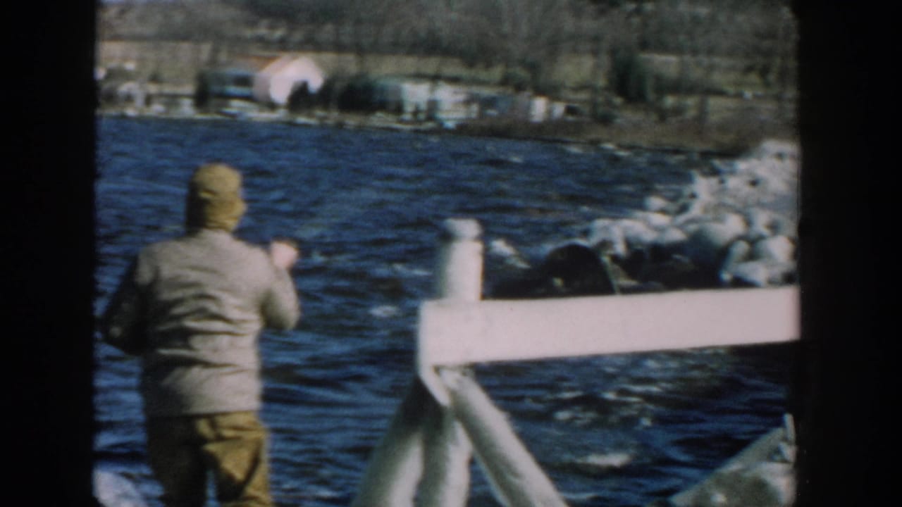 1957 Wisconsin home-movie frame of a man seen from behind on a wooden pier, facing choppy blue river water and a distant shoreline.