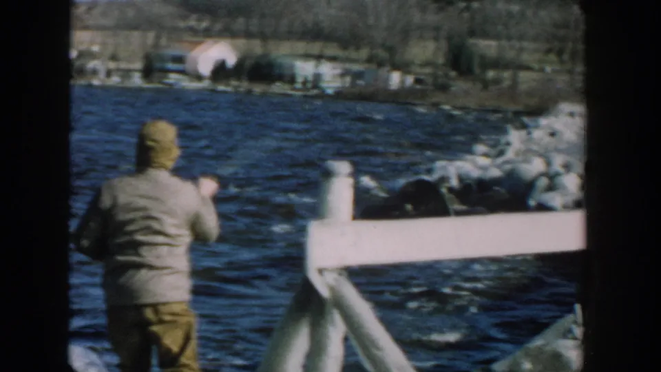 1957 Wisconsin home-movie frame of a man seen from behind on a wooden pier, facing choppy blue river water and a distant shoreline.