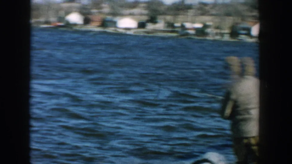 1957 Wisconsin home-movie frame with motion blur as the camera pans past a man fishing beside blue river water.