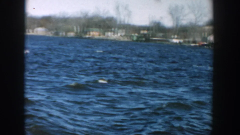 1957 Wisconsin home-movie frame showing open river water, bare shoreline trees, small houses, and distant waterfront structures.