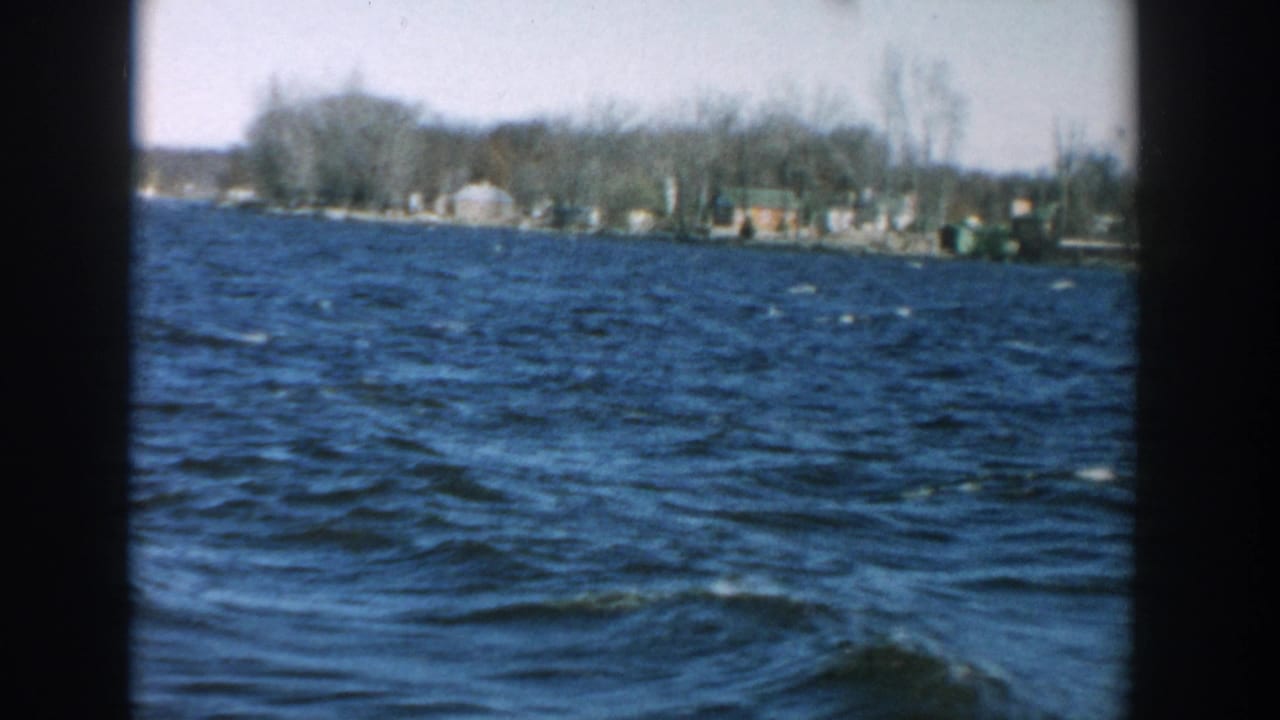 Choppy blue lake water with a distant shoreline of bare trees and small buildings.