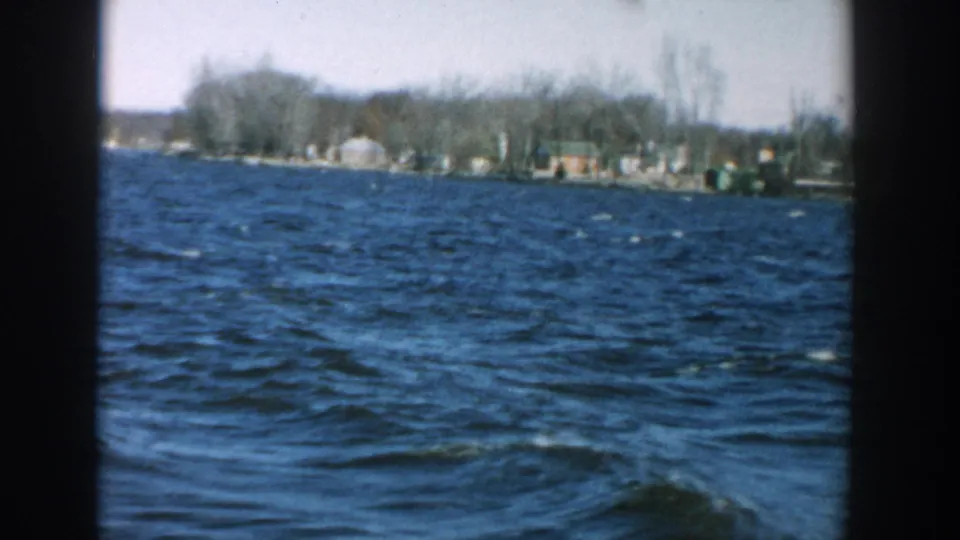 1957 Wisconsin home-movie frame showing rippling lake water and a far shore with bare trees and scattered small buildings.