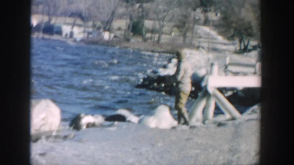 1957 Wisconsin lakeside frame with a person near shoreline rocks, water, and a simple wooden dock or ramp structure.