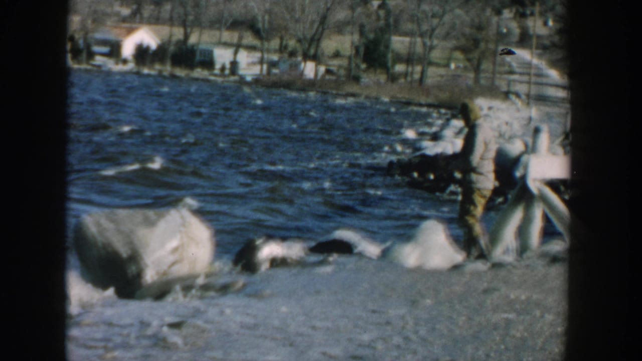 1957 Wisconsin home-movie frame with a man near boulders at the lake edge and a wooded shore visible in the background.