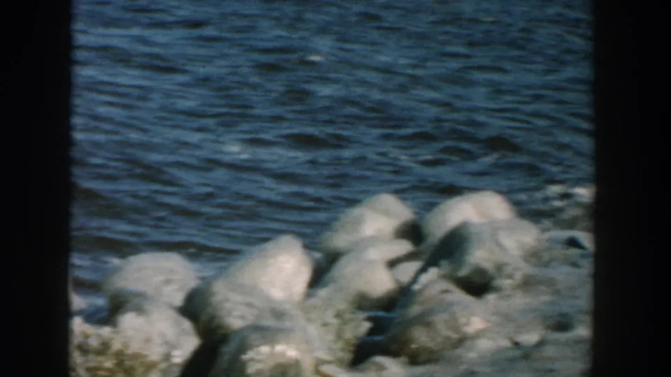 1957 Wisconsin home-movie frame showing choppy lake water and rounded shoreline rocks, with the earlier figure no longer clearly visible.