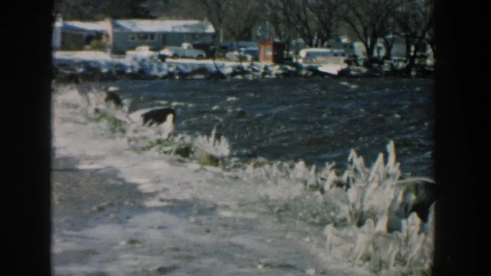 1957 Wisconsin 8mm frame showing icy shore foreground with rough water and a distant lakeside or riverbank area beyond.