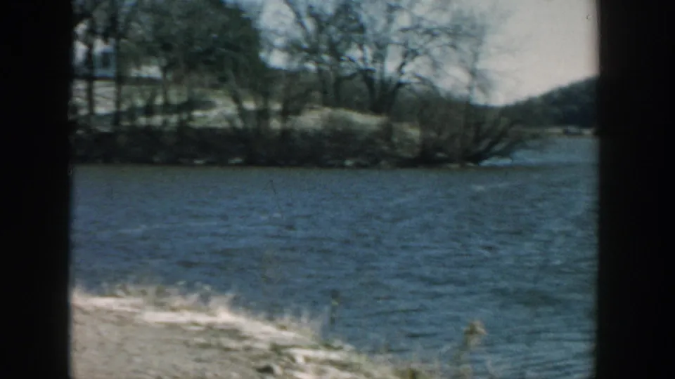 1957 Wisconsin 8mm frame of a quiet lake edge with bare trees, low shoreline, and distant wooded hills.