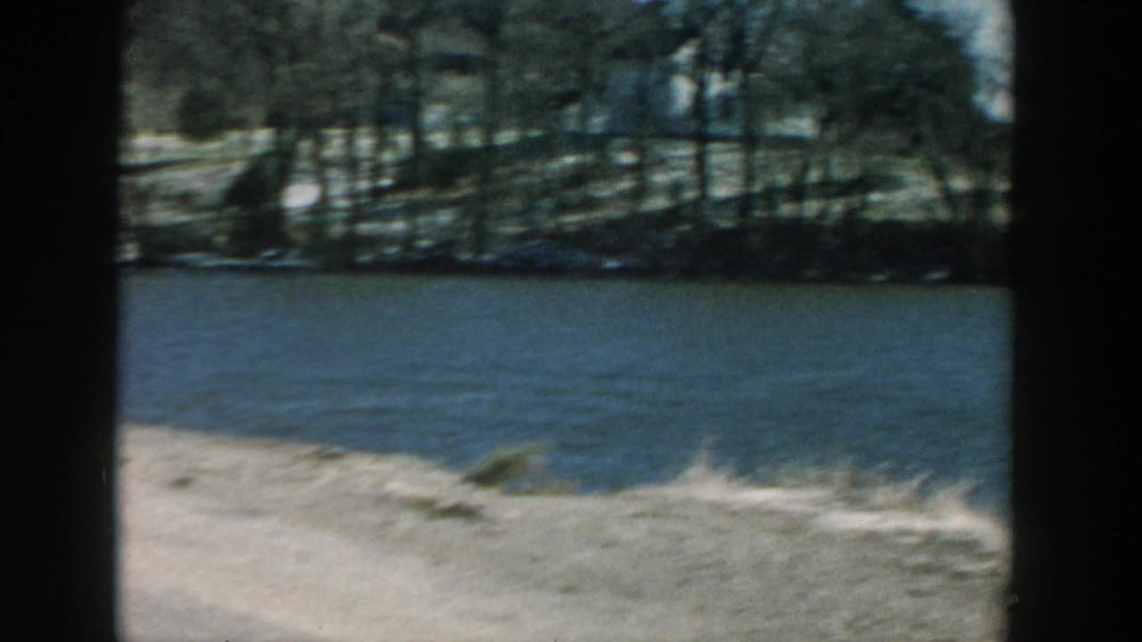 Riverbank and water seen through dark film gate edges from a moving vehicle.