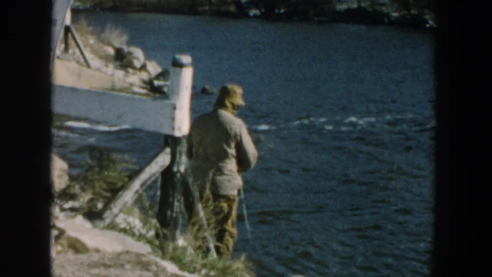 1957 Wisconsin 8mm frame with an older man in outdoor clothing standing by the river and holding a fishing rod.