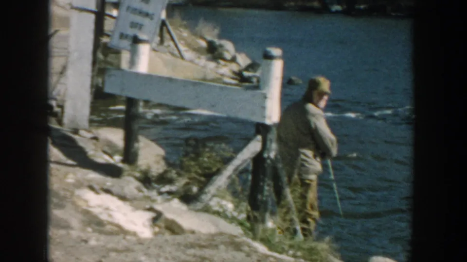 1957 Wisconsin frame showing an older man fishing at the river edge near bridge posts and a small roadside sign.