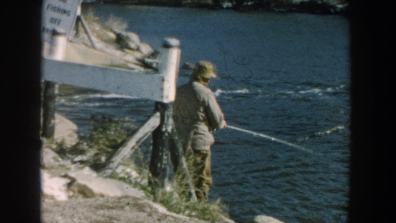 Man fishing beside blue water near a white post on a rocky shoreline.