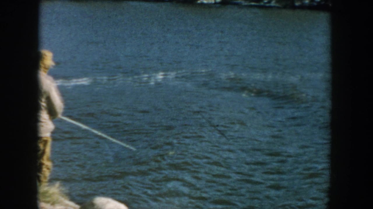 Man in a cap holds a fishing rod toward rippling blue water.