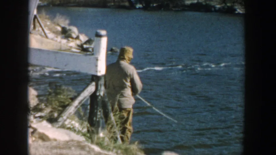 1957 Wisconsin 8mm frame showing a man fishing from a rocky bank, with grasses and water visible around him.