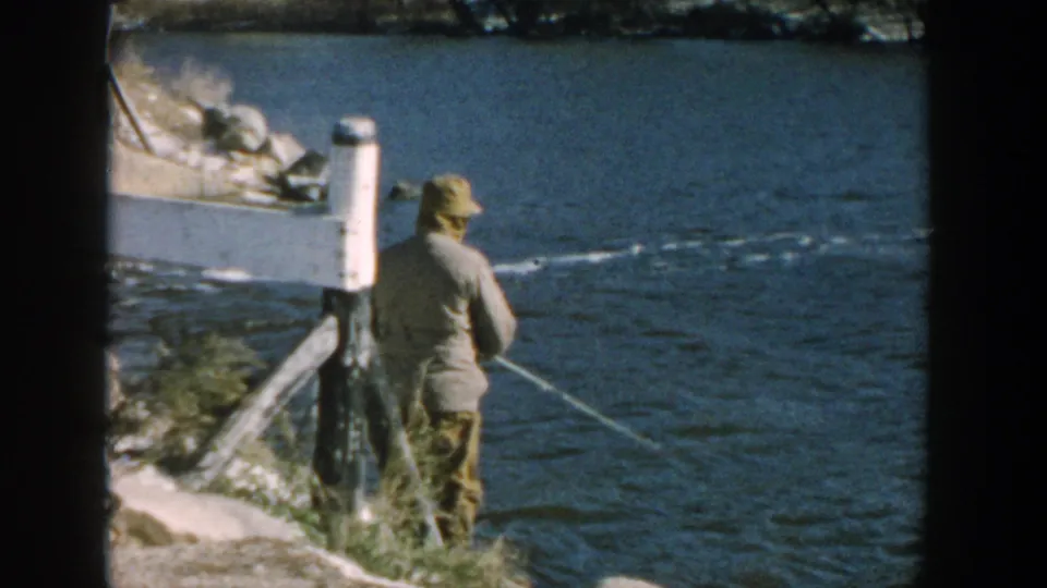 1957 Wisconsin home-movie frame with an adult man in a cap fishing near a white post and rocky shoreline.