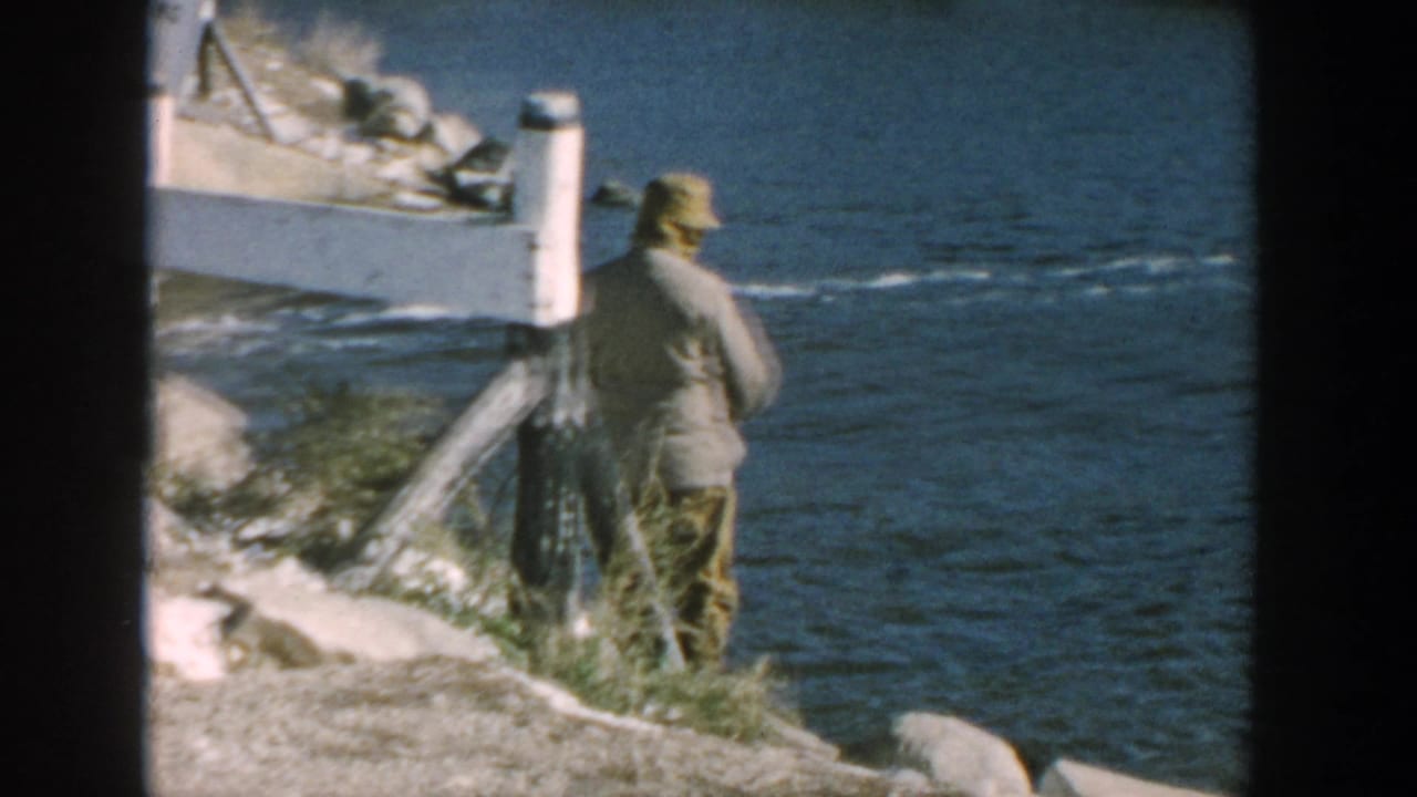 Man in a cap stands on a rocky shoreline beside choppy water.