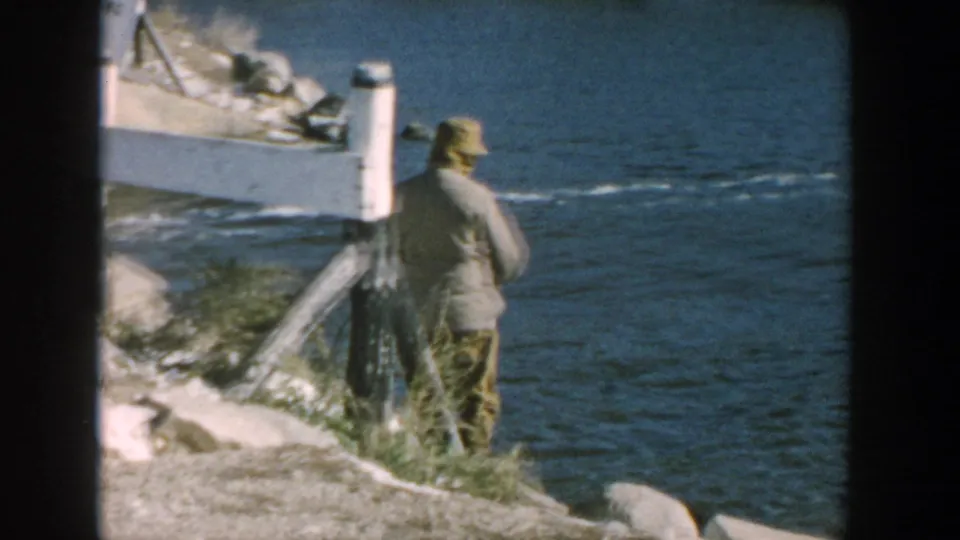 1957 Wisconsin 8mm frame showing a man in a light jacket and cap standing near water on a rocky shore.