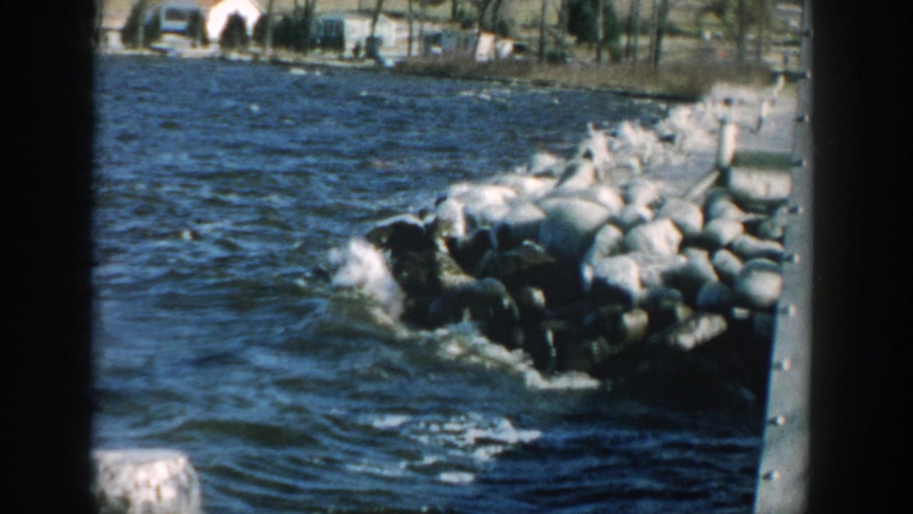 Waves hit a rock jetty with distant shore buildings beyond the water.