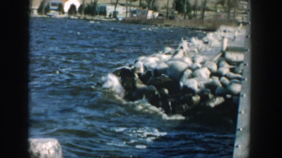 Wisconsin shoreline frame with choppy water breaking against large rocks and distant buildings behind bare trees.