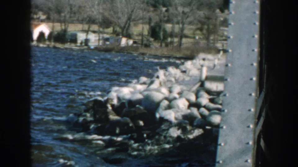 1957 Wisconsin 8mm frame showing waves and spray hitting a rocky shoreline with bare trees and buildings beyond.