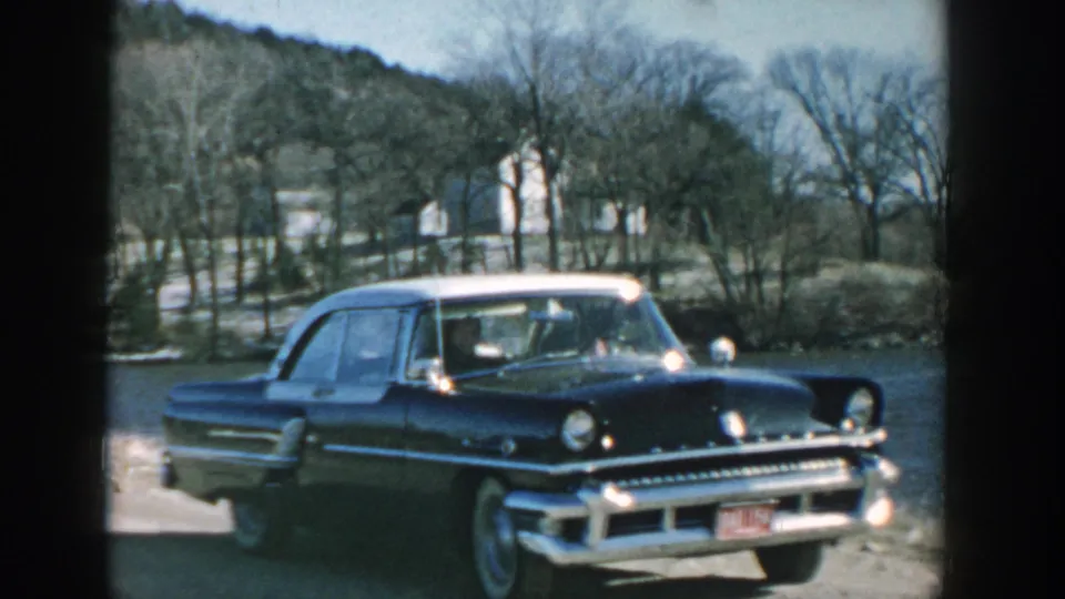 Wisconsin 1957 frame of a dark blue chrome-trim sedan with white roof and whitewall tires beside a river.