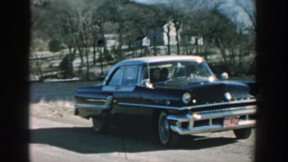 A 1957 Wisconsin home-movie view shows the front of a dark blue mid-century sedan near a river and wooded hillside.