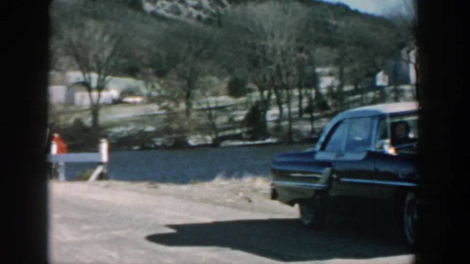 Wisconsin 1957 frame showing the sedan leaving the edge of view, river behind it, and a small distant person near a post.