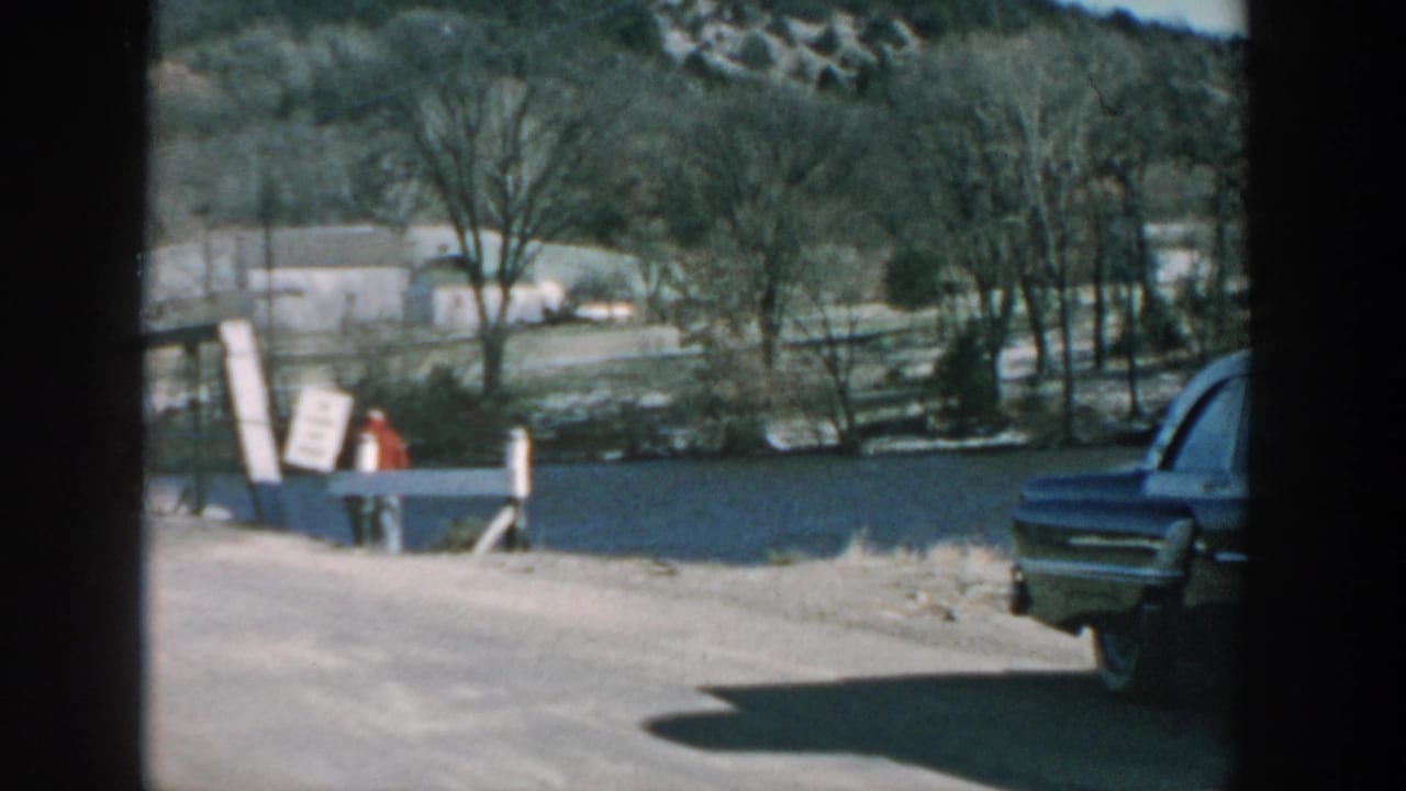 Rural road beside a river with a parked dark sedan near a small wooden bridge.