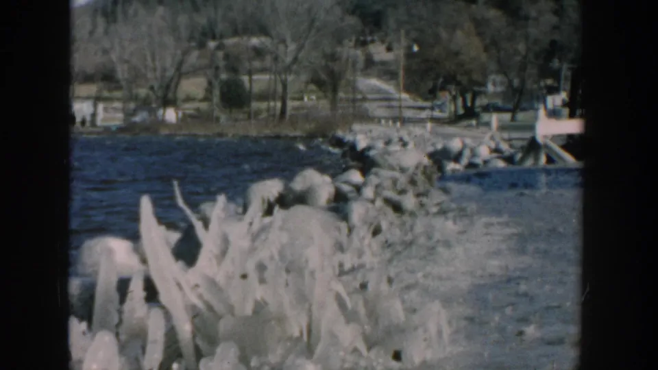 1957 Wisconsin home-movie view of small waves, large boulders, a riverbank, rural road, bridge area, bare trees, and parked vehicle edge.