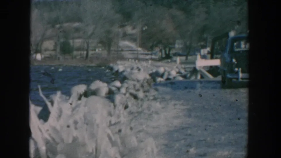 1957 Wisconsin frame showing waves against shoreline rocks, a rural road and bridge area, bare trees, hillside, and a partial dark sedan.