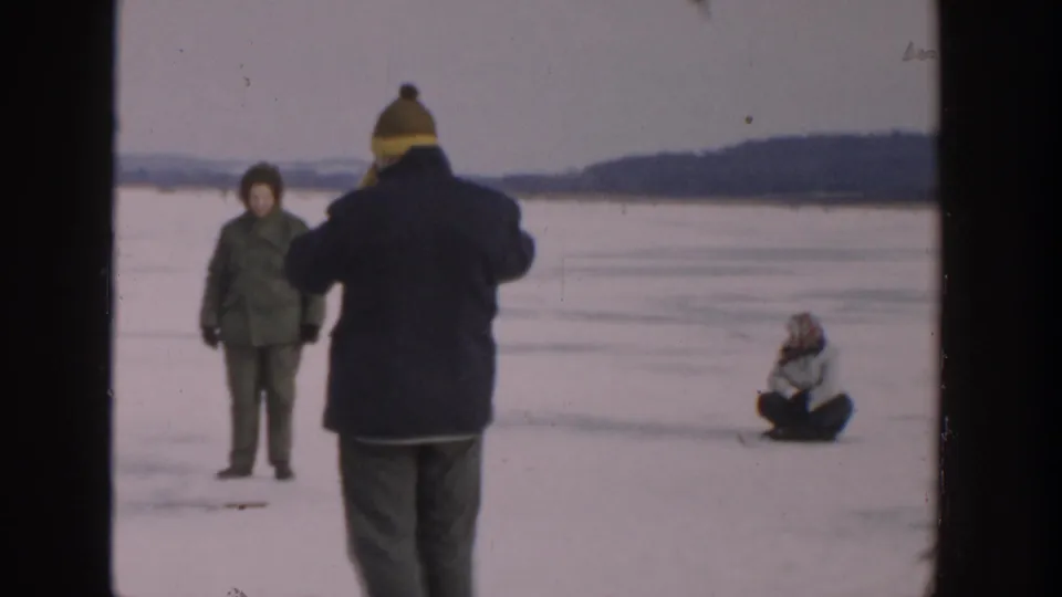 A 1957 Wisconsin winter scene with bundled people on lake ice, including a seated figure and a standing figure facing the camera.