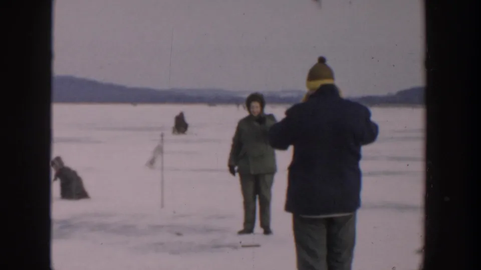 Home-movie frame from Wisconsin in 1957 showing a bundled person near a marker on the ice while others sit farther back.