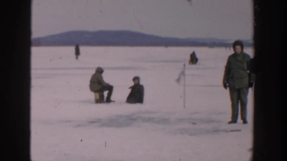 Wide 1957 Wisconsin frozen-lake view with seated ice fishers and a standing bundled person at the right edge.