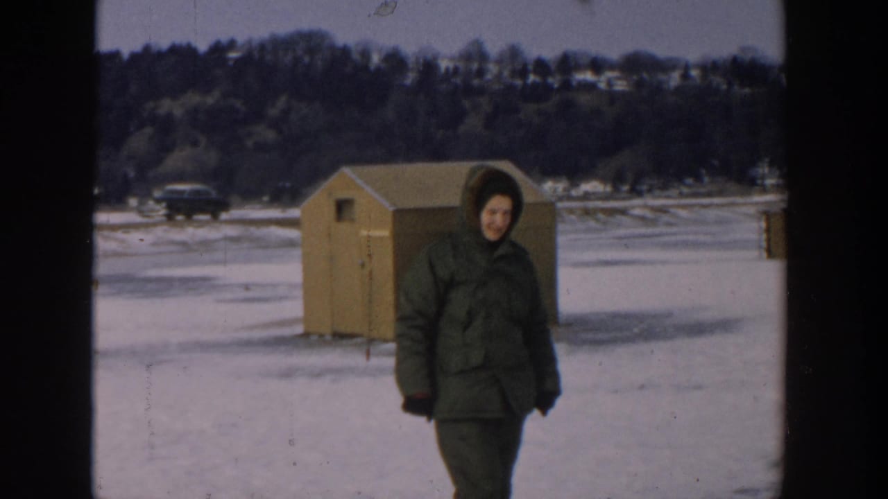 Bundled person walking on a frozen lake near a small wooden shelter.