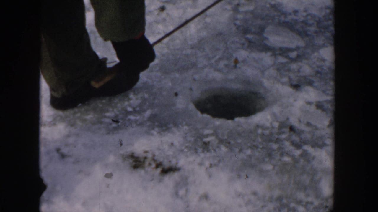A close 1957 Wisconsin view shows a bundled person standing on packed snow beside a round ice-fishing hole and rod.