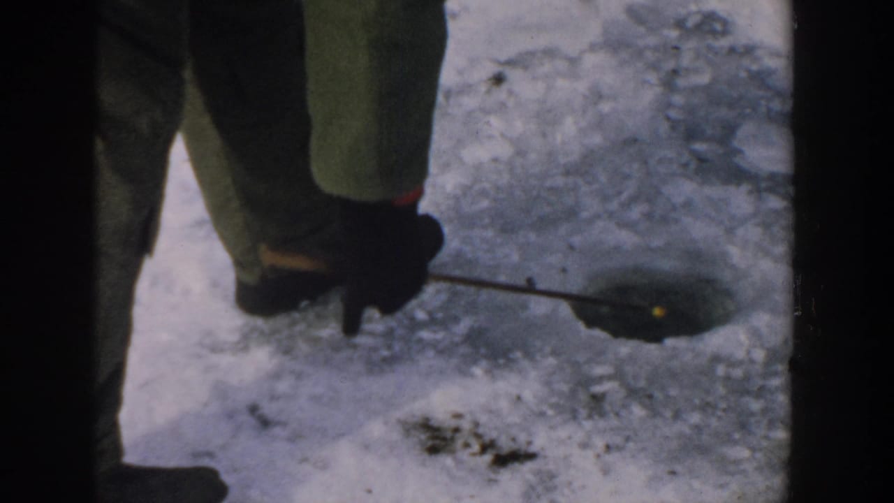 The camera looks down at a bundled person's boots and legs beside an ice-fishing hole and rod on the Wisconsin lake ice.