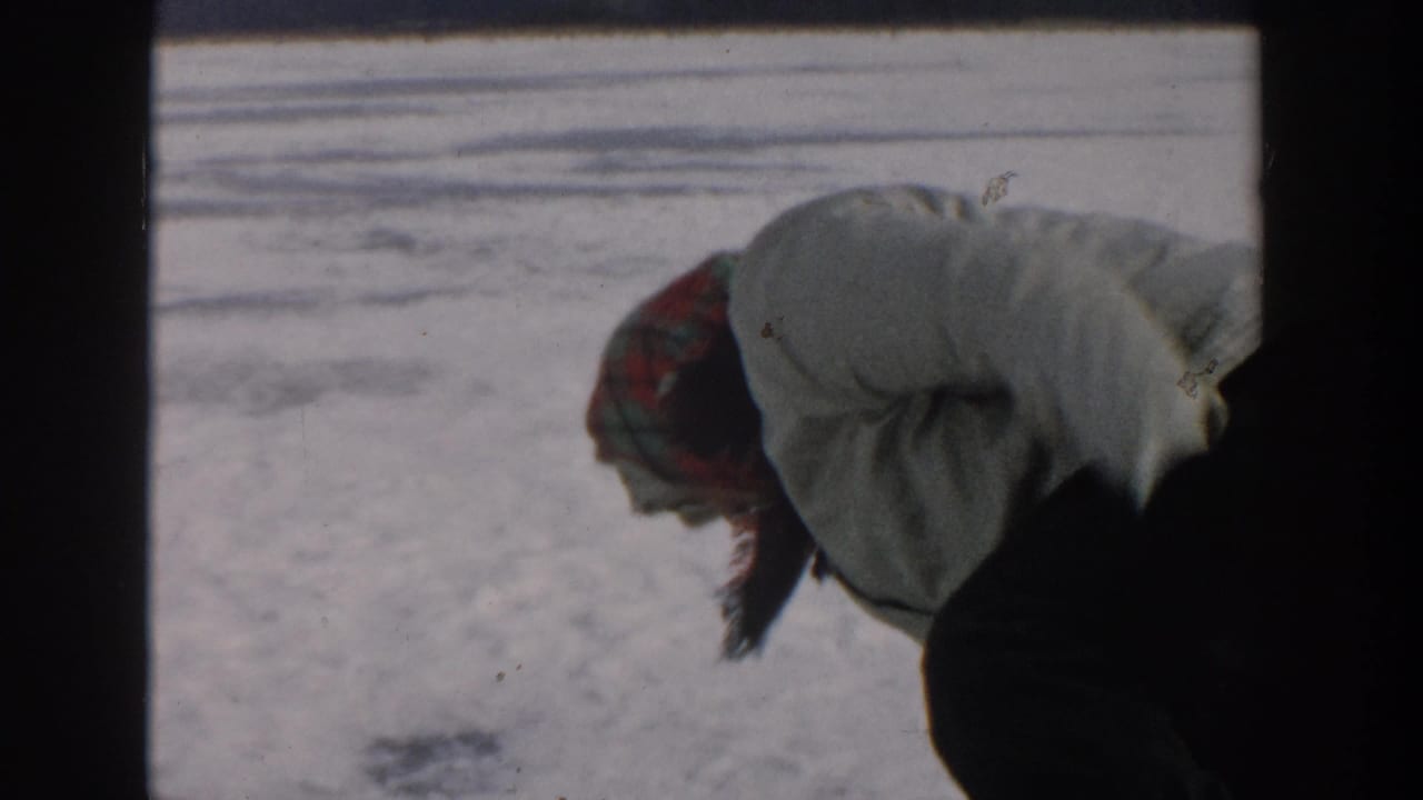 A person in a plaid hat and light jacket bends over the snowy Wisconsin lake ice while tending the fishing spot.