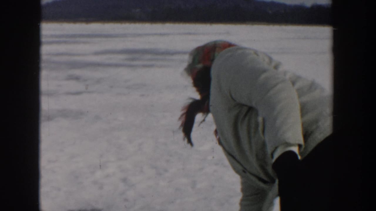 Woman in a light coat bends over snow-covered ice beside a frozen lake.