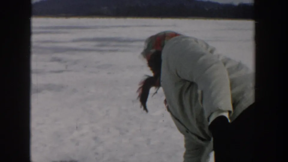 In 1957 Wisconsin home-movie footage, a woman in a light coat and plaid headscarf bends toward the ice on a frozen lake.