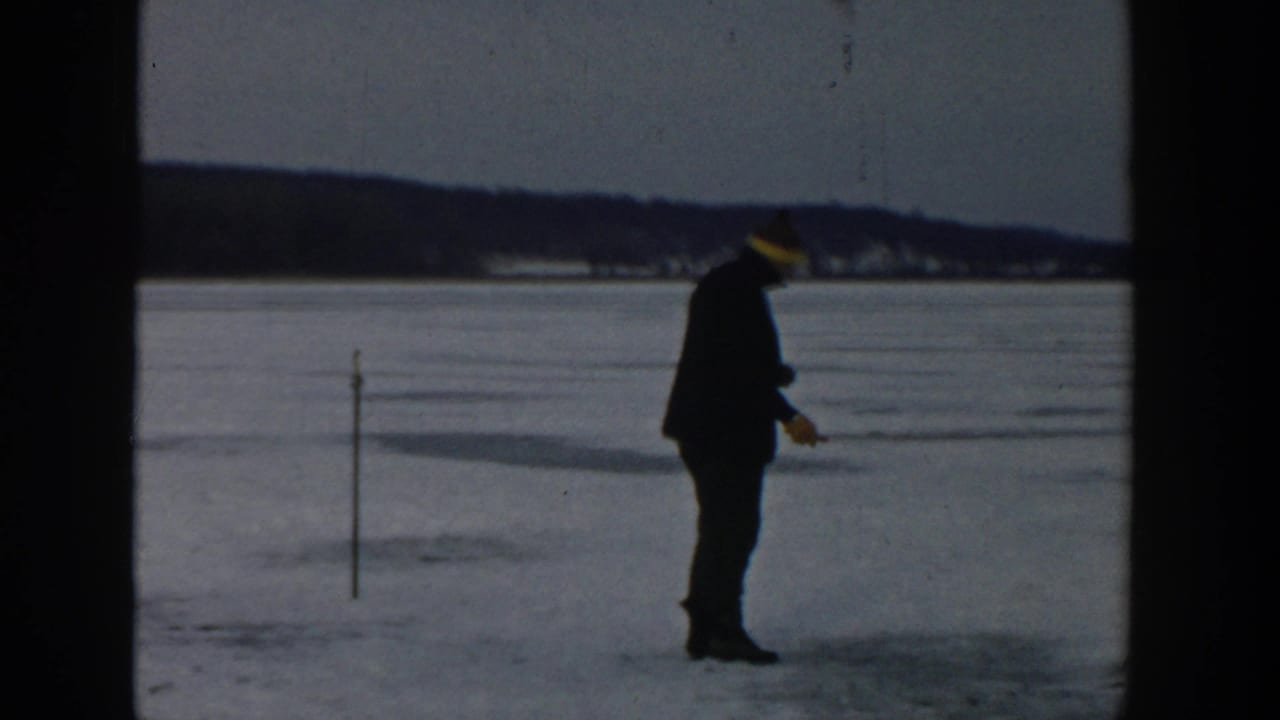 Man in a dark coat and yellow hat stands on a frozen lake near a marker stake.
