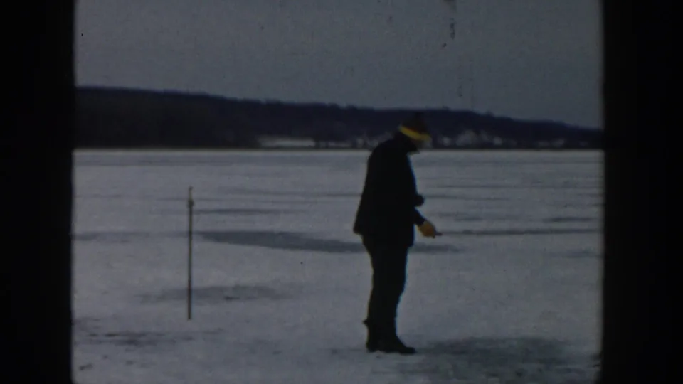 A man in a dark coat and yellow hat stands on the frozen Wisconsin lake near a stake, holding a small tool or object.